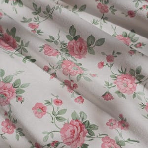 May include: A white fabric featuring a repeating floral pattern of pink roses with sage green leaves and stems. The material is draped in soft folds, creating a textured surface that highlights the botanical print across the textile.
