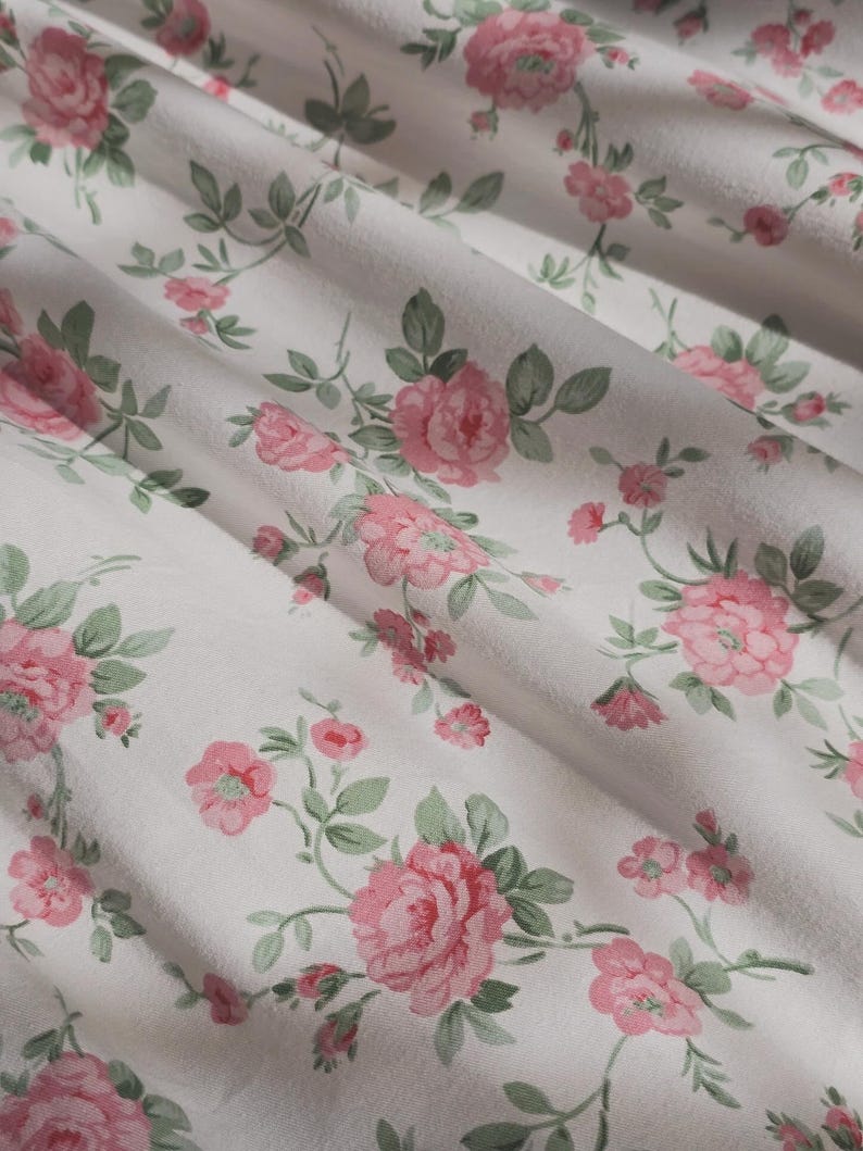 May include: A white fabric featuring a repeating floral pattern of pink roses with sage green leaves and stems. The material is draped in soft folds, creating a textured surface that highlights the botanical print across the textile.