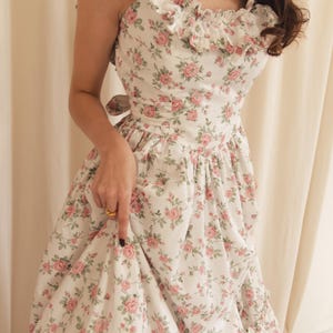 May include: A white sleeveless dress featuring a repeating pattern of pink roses and green leaves. The dress has a square neckline with ruffled trim along the shoulders and bodice, a fitted waist with a tie-back detail, and a full, gathered skirt.