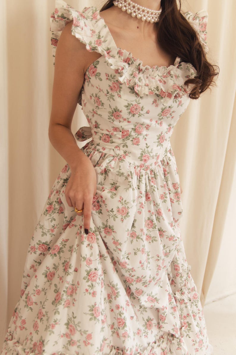 May include: A white sleeveless dress featuring a repeating pattern of pink roses and green leaves. The dress has a square neckline with ruffled trim along the shoulders and bodice, a fitted waist with a tie-back detail, and a full, gathered skirt.