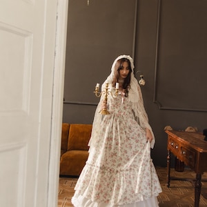 May include: A floor-length dress featuring a cream base with a pink and green floral pattern, a tiered skirt, and a ruffled hem. The garment is paired with a sheer, lace-trimmed head covering and a gold-toned metal candelabra with three candle holders.