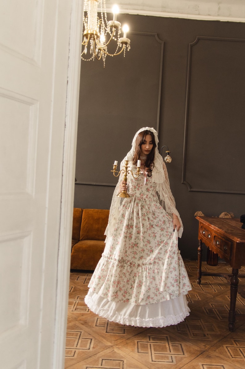 May include: A floor-length dress featuring a cream base with a pink and green floral pattern, a tiered skirt, and a ruffled hem. The garment is paired with a sheer, lace-trimmed head covering and a gold-toned metal candelabra with three candle holders.