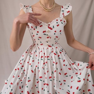 K&ouml;nnte beinhalten: Wei&szlig;es Kleid mit rotem Kirschmuster. Es hat einen herzf&ouml;rmigen Ausschnitt, eine Knopfleiste vorne und R&uuml;schentr&auml;ger. Der volle Rock erzeugt eine Vintage-inspirierte Silhouette.