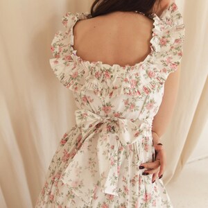 May include: A white dress featuring a repeating pattern of small pink roses and green leaves. The garment has a square neckline with wide ruffled trim along the shoulders and a large fabric bow tied at the center of the back waistline.