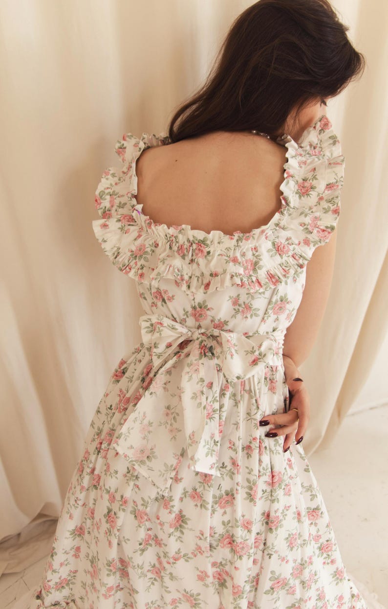 May include: A white dress featuring a repeating pattern of small pink roses and green leaves. The garment has a square neckline with wide ruffled trim along the shoulders and a large fabric bow tied at the center of the back waistline.