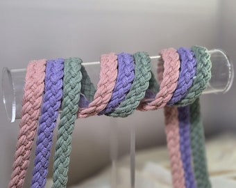 Op de afbeelding: Gevlochten katoenen koorden in pastelkleuren roze, lavendel en groen worden getoond op een heldere acryl staaf. De koorden zijn strak geweven, met een textuurpatroon. De presentatie benadrukt de kleuren en textuur van de koorden.