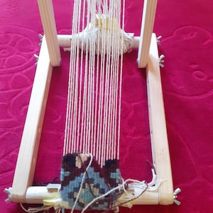 Puede incluir: Un telar de madera con una urdimbre de hilo blanco y un tapiz tejido de colores en la parte inferior. El telar est&aacute; sobre una superficie roja.