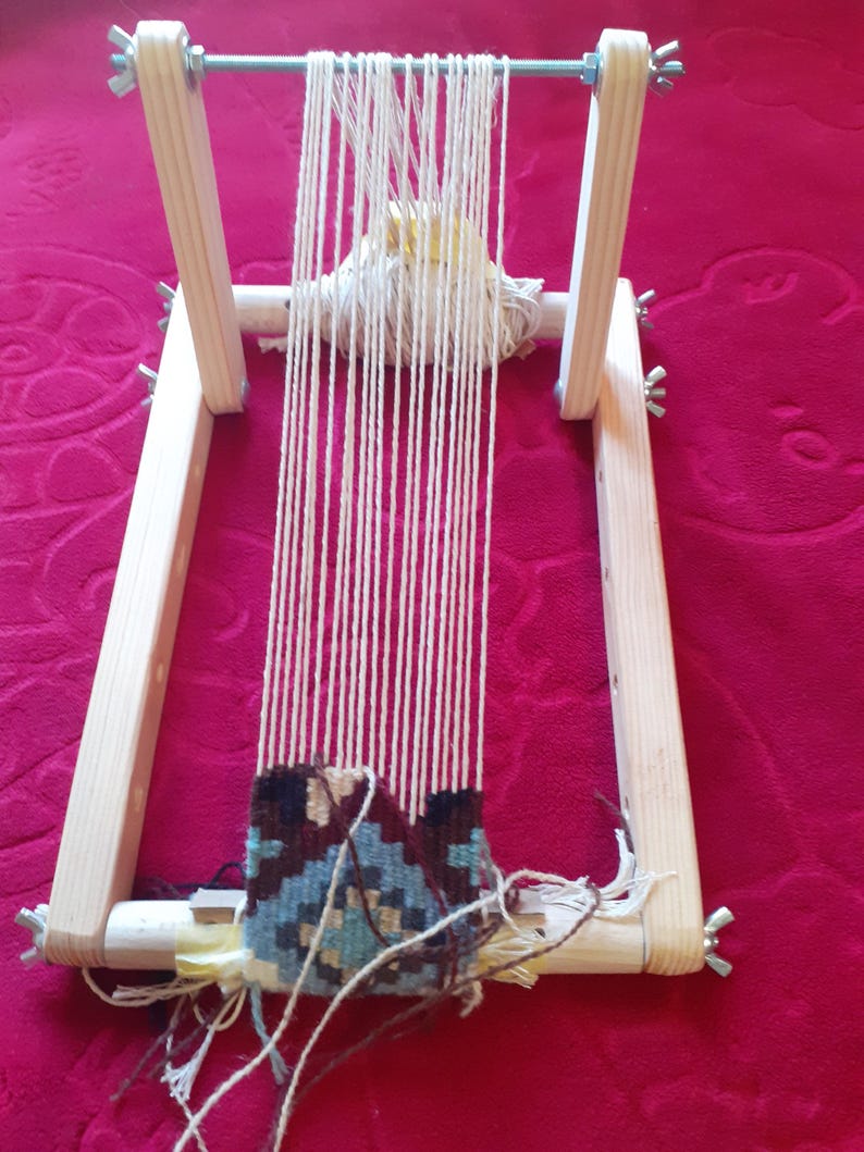 Puede incluir: Un telar de madera con una urdimbre de hilo blanco y un tapiz tejido de colores en la parte inferior. El telar est&aacute; sobre una superficie roja.