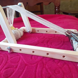 Puede incluir: Un telar de madera con una urdimbre de hilo blanco y un tapiz parcialmente tejido en azul, marr&oacute;n y blanco. El telar tiene un rodillo de madera para enrollar el hilo y un marco de madera con tornillos de metal.