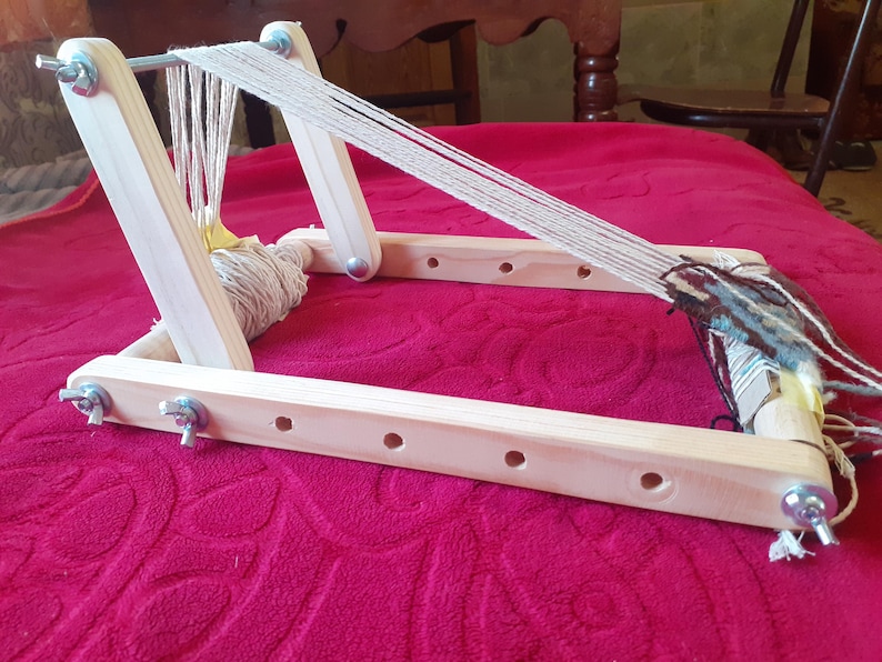 Puede incluir: Un telar de madera con una urdimbre de hilo blanco y un tapiz parcialmente tejido en azul, marr&oacute;n y blanco. El telar tiene un rodillo de madera para enrollar el hilo y un marco de madera con tornillos de metal.
