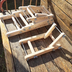 Könnte beinhalten: Zwei Holzwebstühle, einer ist zusammengeklappt und der andere ist offen. Beide Webstühle sind aus Naturholz gefertigt und haben Metallbeschläge.