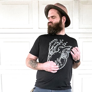 May include: A man wearing a black t-shirt with a white graphic of a heart with a guitar inside. He is wearing a brown fedora and blue jeans.