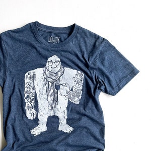 Op de afbeelding: Een donkerblauw T-shirt met een witte afbeelding van een yeti die een kopje koffie vasthoudt. De yeti heeft tatoeages op zijn armen en draagt een sjaal. De tekst "DOWDY" is op het shirt gedrukt.