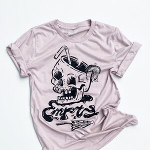 May include: A light pink t-shirt with a black graphic of a skull with a straw in it and the word "Empty" written below it.