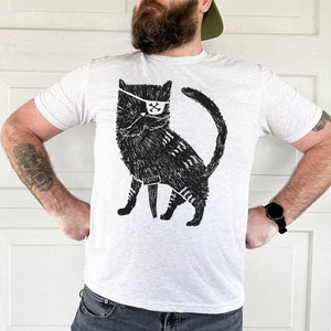 以下が含まれることがあります： 黒い眼帯と剣を持った猫の白黒イラストが描かれた白いTシャツ。猫は後ろ足で立ち、獰猛な表情をしている。