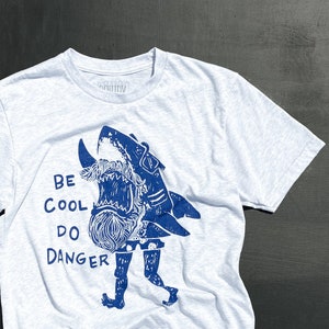 May include: A white t-shirt with a blue graphic of a shark with a beard and the text "Be Cool Do Danger".