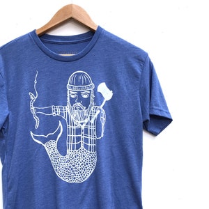 Op de afbeelding: Blauw t-shirt met een witte afbeelding van een bebaarde man met een zeemeerminstaart, die een bijl en een slang vasthoudt. De man draagt een muts en een geruit overhemd. Het t-shirt hangt aan een houten hanger.