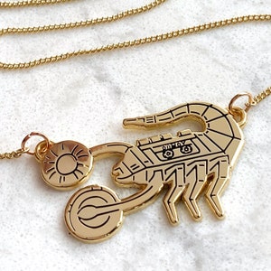 May include: Gold necklace with a scorpion pendant. The scorpion is detailed with a small car on its back. The necklace is on a white surface.