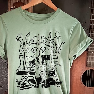 May include: A light green t-shirt with a black and white graphic print of a three-headed monster playing musical instruments. The monster has horns, fangs, and three eyes. The monster is playing a keyboard, drums, and a trumpet.