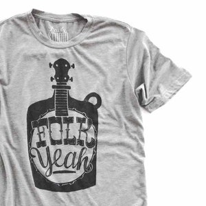 Op de afbeelding: Een grijs T-shirt met een zwart-witte afbeelding van een banjo met de tekst "Folk Yeah" erop.