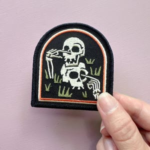 May include: Embroidered patch with a white skeleton design within a black, arched frame. The skeleton is in a graveyard scene with green grass and an orange border. The patch has a black border.