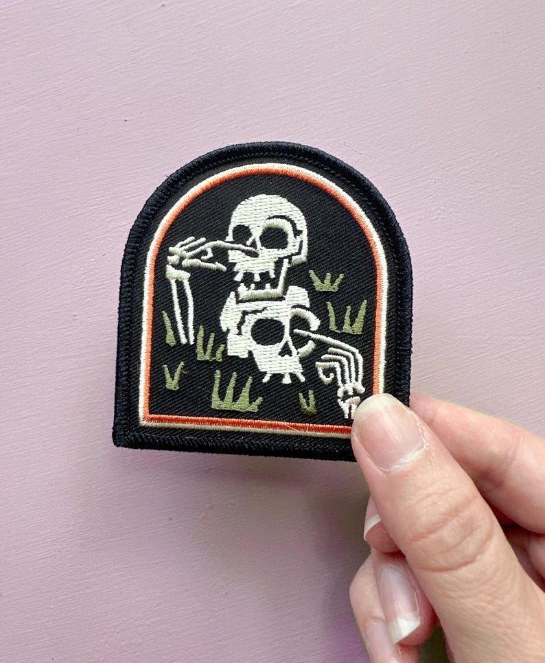 May include: Embroidered patch featuring a white skeleton in a black arch-shaped frame. The skeleton is in a graveyard setting with green grass and an orange border. The patch has a black border.