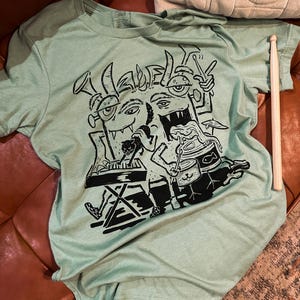 Op de afbeelding: Een lichtgroen T-shirt met een zwart-witte grafische print van een band cartoonmonsters die instrumenten bespelen. Het monster in het midden bespeelt een keyboard, het monster links bespeelt een trompet en het monster rechts bespeelt een drumstel.