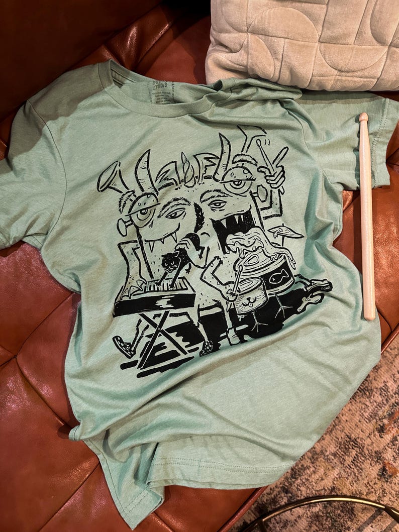 Op de afbeelding: Een lichtgroen T-shirt met een zwart-witte grafische print van een band cartoonmonsters die instrumenten bespelen. Het monster in het midden bespeelt een keyboard, het monster links bespeelt een trompet en het monster rechts bespeelt een drumstel.