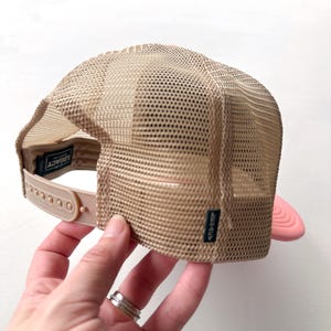 May include: A tan trucker hat with a mesh back and a pink peak. The hat features a snap closure and a small label that reads "LEGACY". The hat is being held.