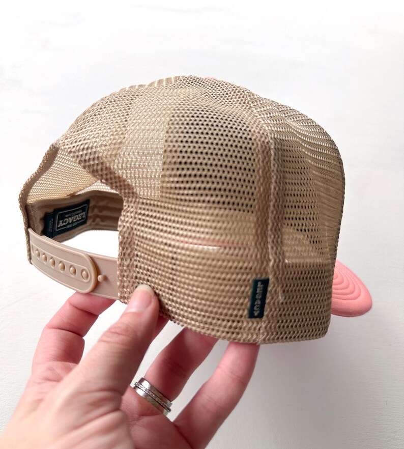 May include: A tan trucker hat with a mesh back and a pink peak. The hat features a snap closure and a small label that reads "LEGACY". The hat is being held.