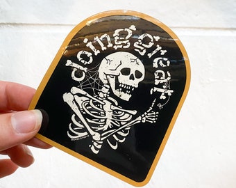 May include: A black and white sticker with a skeleton holding a bone and a spider web. The sticker has a gold border and the text "doing great" written around the skeleton.