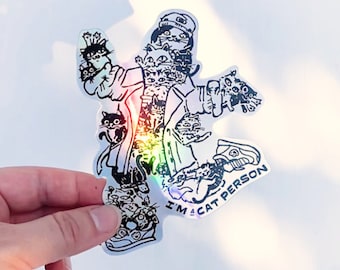 May include: Holographic sticker featuring a cartoon illustration of a person composed of cats. The design includes a hat, sneakers, and the text "I'M A CAT PERSON". The sticker has a rainbow sheen and is held by a hand.