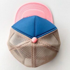 May include: A blue and pink trucker hat with a tan mesh back. The hat has a pink button on the front. This casual hat is a stylish accessory. The hat's design is suitable for various outdoor activities.