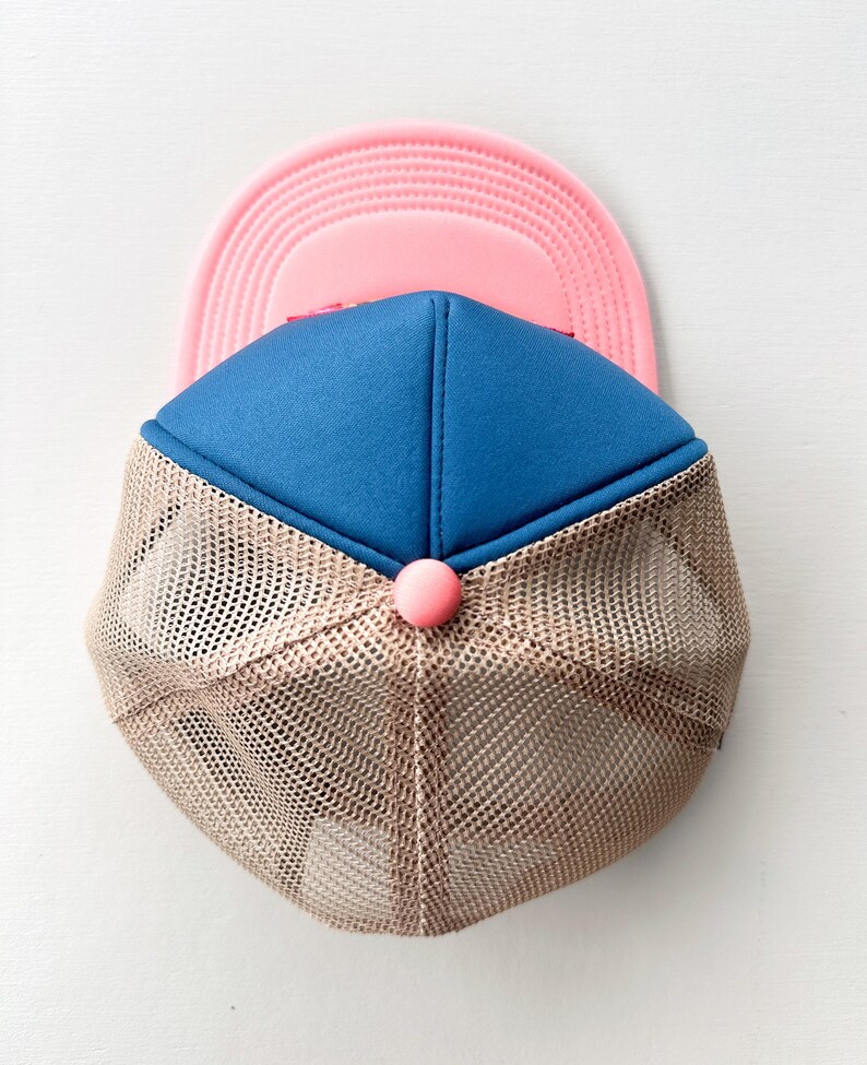 May include: A blue and pink trucker hat with a tan mesh back. The hat has a pink button on the front. This casual hat is a stylish accessory. The hat's design is suitable for various outdoor activities.