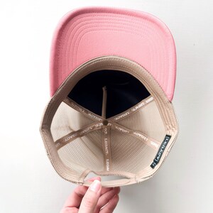 May include: A pink trucker hat with a mesh back and a curved brim. The interior features a navy blue front panel and a tan mesh. The hat has the word "LEGACY" printed on the interior.