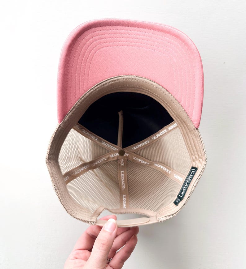 May include: A pink trucker hat with a mesh back and a curved brim. The interior features a navy blue front panel and a tan mesh. The hat has the word "LEGACY" printed on the interior.