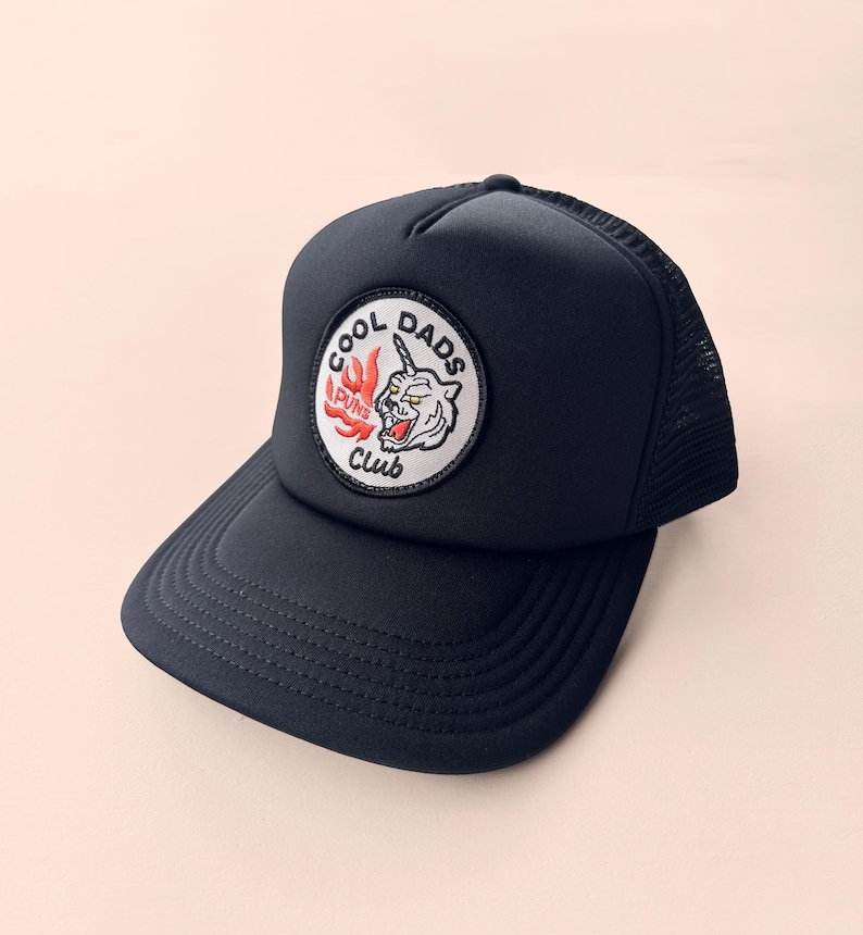 May include: Navy blue trucker hat with a curved peak and mesh back. The hat features a circular patch with the text "COOL DADS CLUB" surrounding a tiger illustration with flames. A stylish accessory.