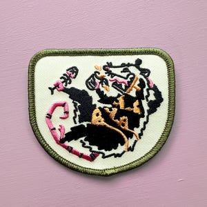 May include: Embroidered patch featuring a stylized black and tan animal with a pink and white bone. The patch has a cream-colored background with a green border. The patch is shield-shaped.
