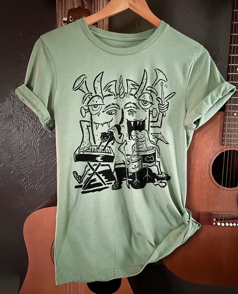 Op de afbeelding: Een lichtgroen T-shirt met een zwart-witte grafische print van een driekoppig monster dat muziekinstrumenten bespeelt. Het monster heeft hoorns, tanden en drie ogen. Het monster bespeelt een keyboard, drums en een trompet.