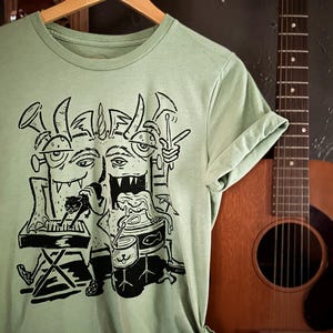 Op de afbeelding: Een lichtgroen T-shirt met een zwart-witte illustratie van een meerkoppig monster dat muziekinstrumenten bespeelt. Het monster speelt keyboard, drums en gitaar en houdt een microfoon vast.