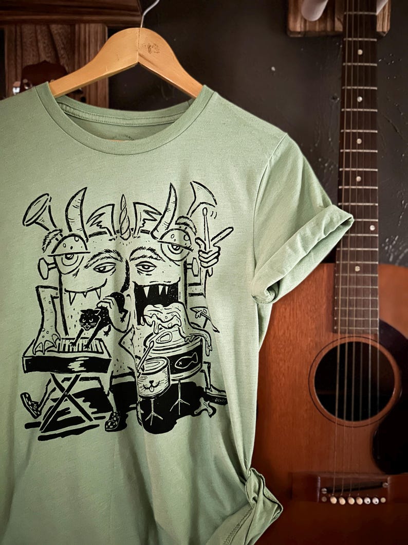 Op de afbeelding: Een lichtgroen T-shirt met een zwart-witte illustratie van een meerkoppig monster dat muziekinstrumenten bespeelt. Het monster speelt keyboard, drums en gitaar en houdt een microfoon vast.