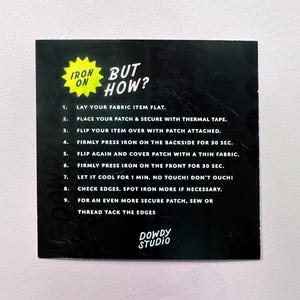 May include: A black instruction card with the text "IRON ON BUT HOW?" and numbered steps for applying a patch. The card features a yellow starburst graphic and the "DOWDY STUDIO" logo.