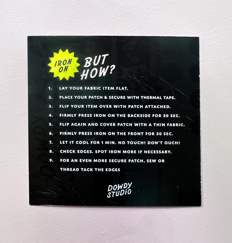 May include: A black instruction card with the text "IRON ON BUT HOW?" and numbered steps for applying a patch. The card features a yellow starburst graphic and the "DOWDY STUDIO" logo.