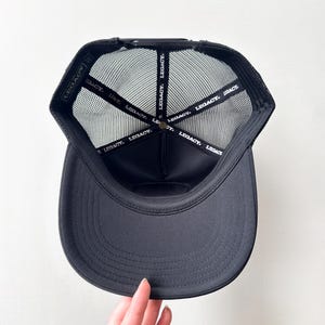 May include: Black trucker hat with a curved peak and mesh back. The interior features a black band with the word "LEGACY" printed repeatedly. The hat is viewed from the inside, showcasing the brand's logo.