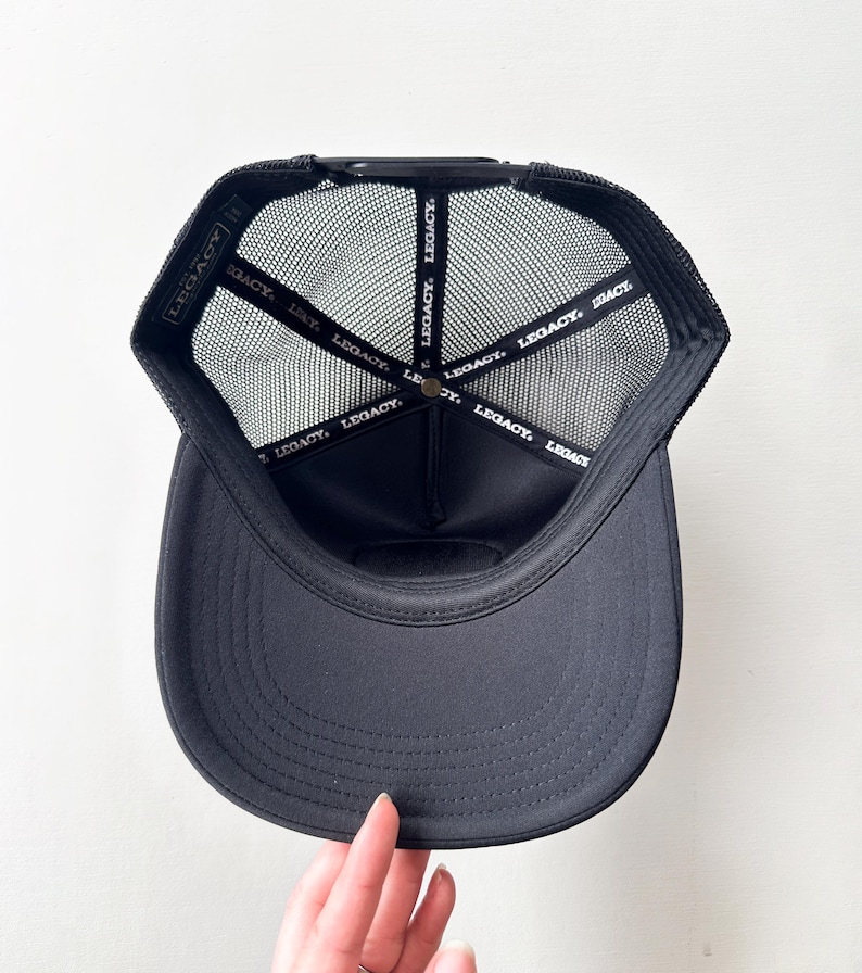 May include: Black trucker hat with a curved peak and mesh back. The interior features a black band with the word "LEGACY" printed repeatedly. The hat is viewed from the inside, showcasing the brand's logo.