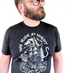 May include: A dark grey t-shirt featuring a white graphic of an opossum riding a bicycle. The graphic includes the text "We Ride At Night" and "Be Goneish by Dawnish".