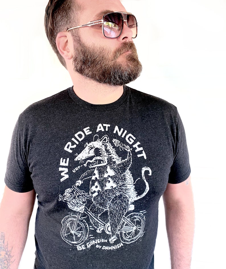 May include: A dark grey t-shirt featuring a white graphic of an opossum riding a bicycle. The graphic includes the text "We Ride At Night" and "Be Goneish by Dawnish".