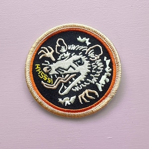 May include: Embroidered patch with a stylized opossum head, open-mouthed and with its tongue out. The opossum is white with black details, set against a black background. The patch has an orange border and the word "MASSS" in yellow.