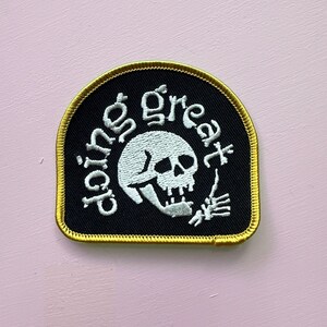 Puede incluir: Parche negro con borde dorado que presenta una calavera blanca y las palabras "doing great" en escritura blanca. La calavera está haciendo una señal de pulgar hacia arriba con una mano de esqueleto. El parche está sobre un fondo rosa claro.