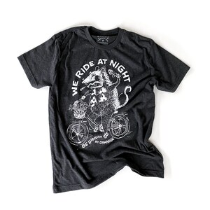 May include: A dark grey t-shirt with a white graphic of a possum riding a bicycle. The graphic says "We Ride At Night" and "Be Goneish By Dawnish".